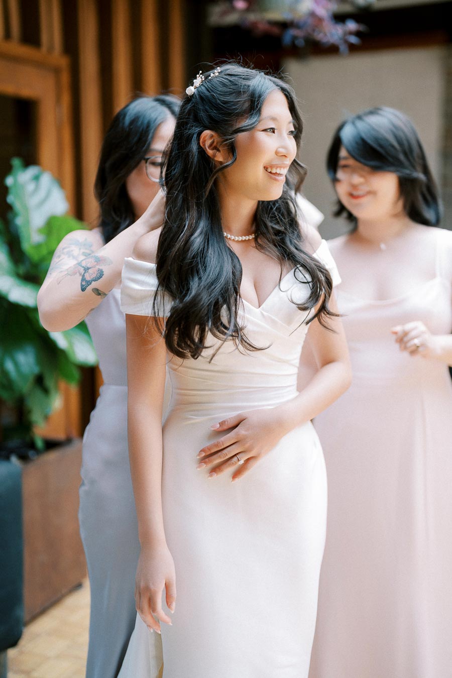 Bride in elegant white dress surrounded by bridesmaids adjusting her gown, smiling and preparing for wedding ceremony.