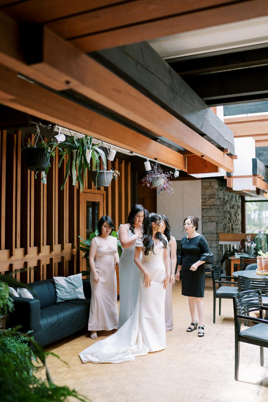 Bride in elegant white gown being assisted by bridesmaids in pastel dresses and a woman in black, preparing for wedding in a beautifully decorated indoor venue with wooden beams and plants.