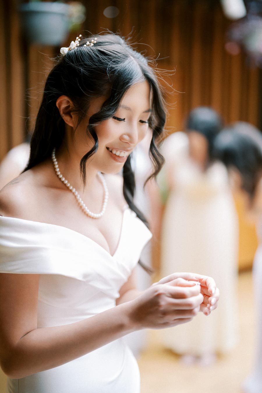 Smiling bride in an elegant white wedding dress and pearl necklace, fiddling with her fingers indoors.