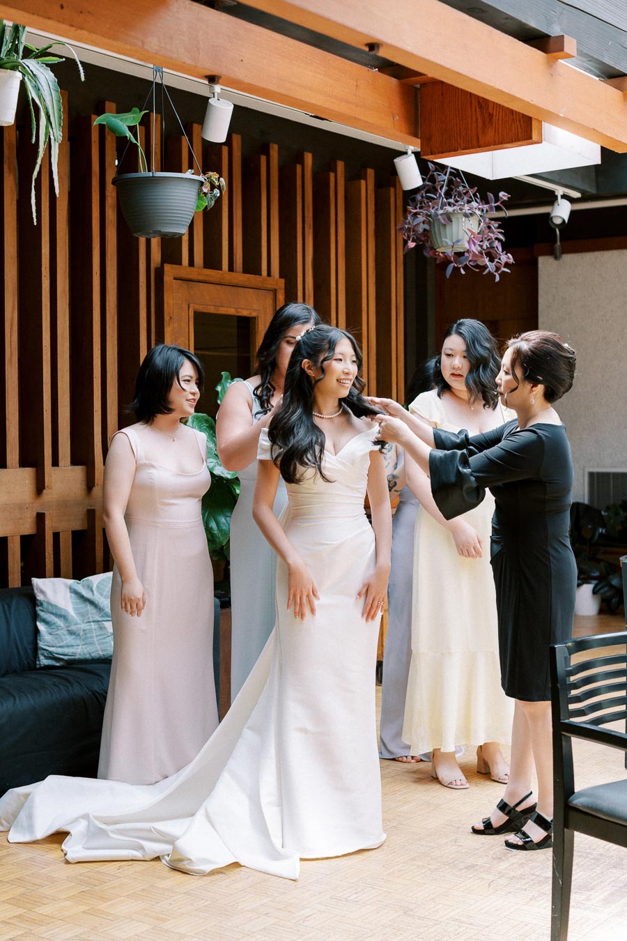 Bridal party assisting bride with gown and jewelry before wedding ceremony in elegantly decorated room.