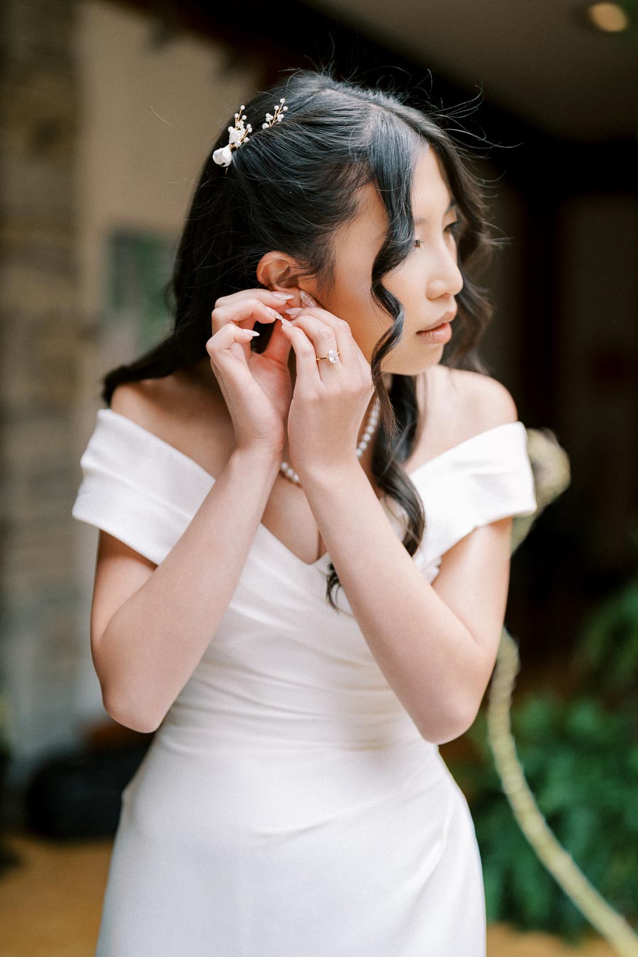 A bride in an elegant off-the-shoulder wedding dress adjusts her earring while preparing for her wedding day.