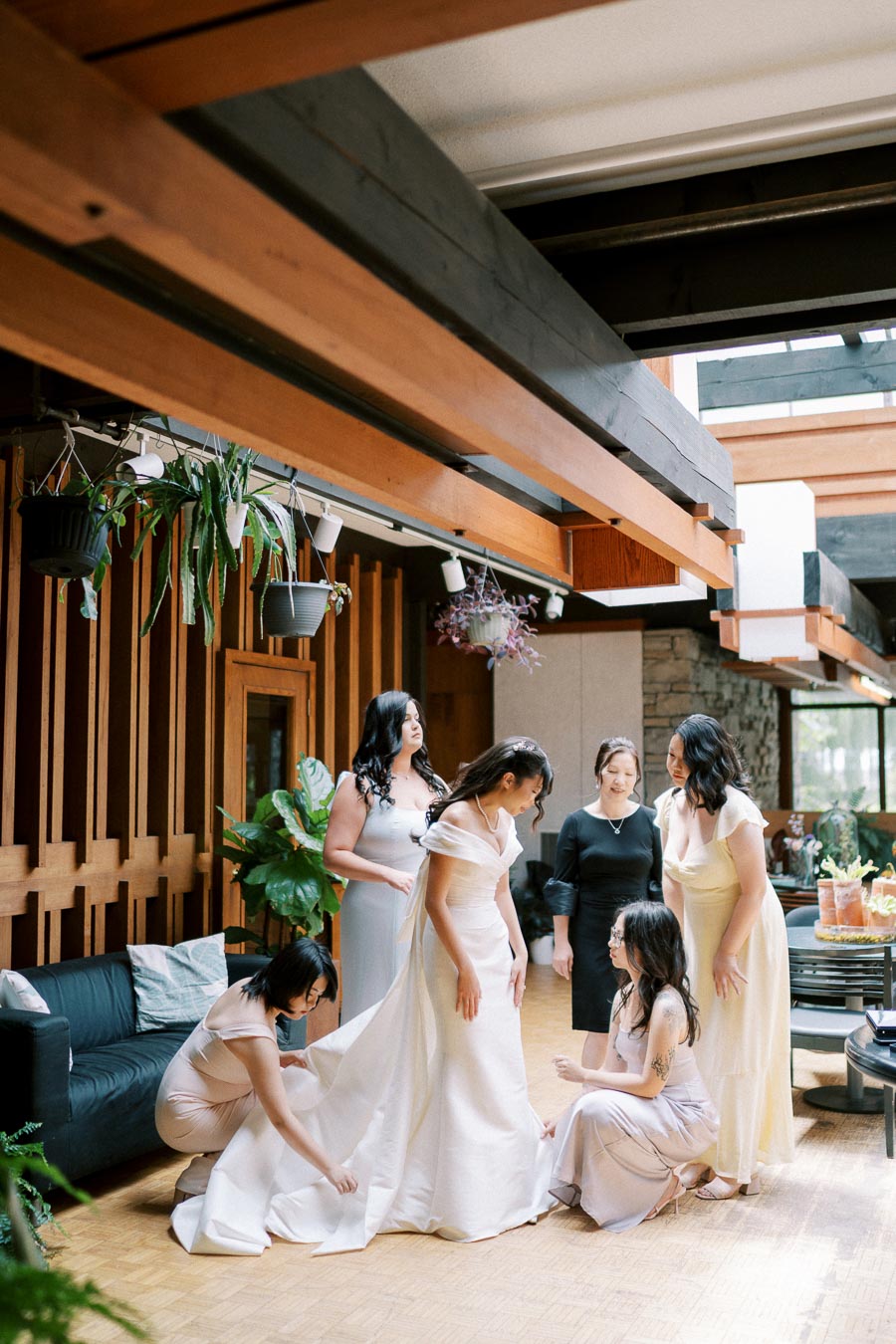 Bridesmaids helping bride adjust her gown in a modern indoor garden setting, highlighting joyful wedding preparations.