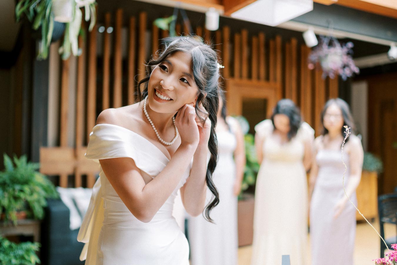 Smiling bride wearing pearl jewelry adjusts earring while standing in a modern, plant-filled room with bridesmaids in elegant dresses blurred in the background.