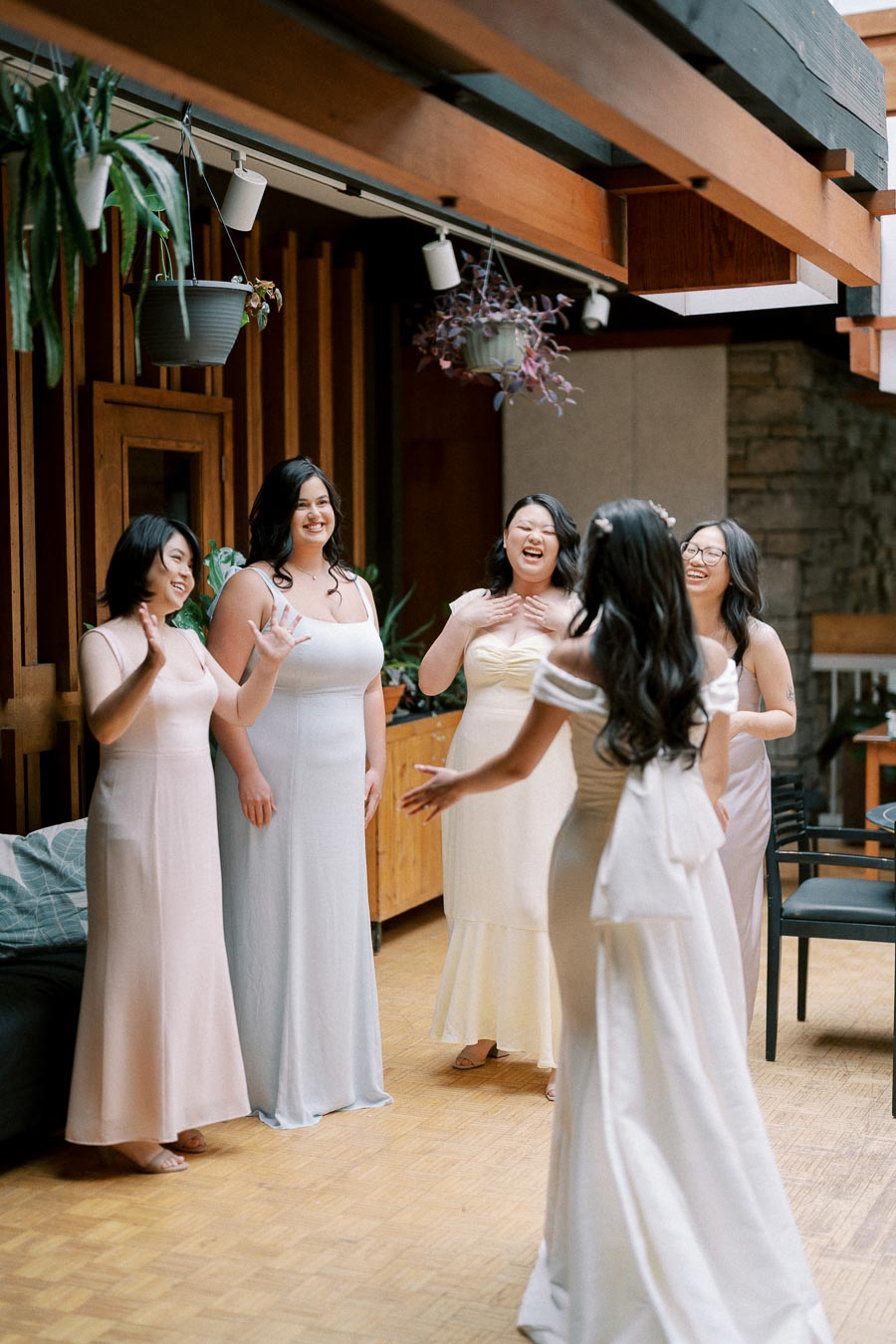 Bride in an elegant white gown joyfully greets a group of bridesmaids in pastel dresses, surrounded by wooden decor and hanging plants, creating a warm and celebratory atmosphere.