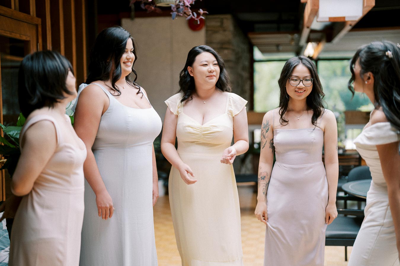 Group of five women in elegant dresses smiling and engaged in conversation in an indoor setting, highlighting friendship and celebration.