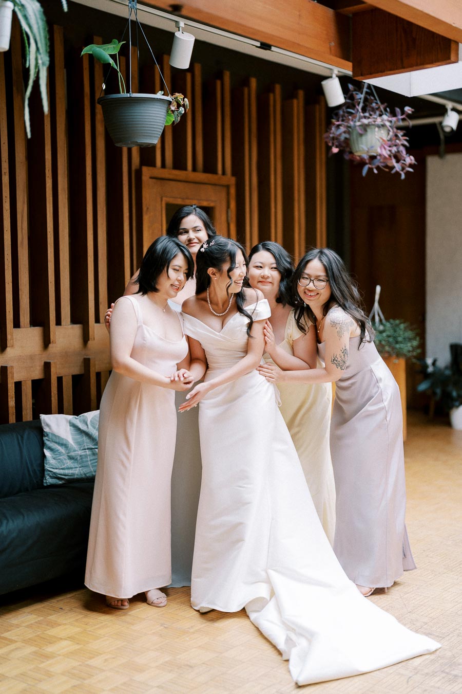 A bride in a white wedding dress joyfully poses with her bridesmaids, who are wearing pastel dresses, in a modern setting with wooden paneling and hanging plants.