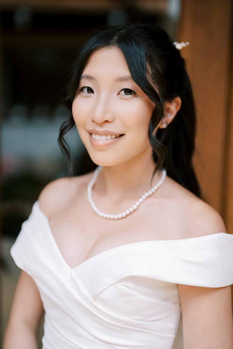 Bride smiling in an elegant off-shoulder white wedding gown with a pearl necklace.