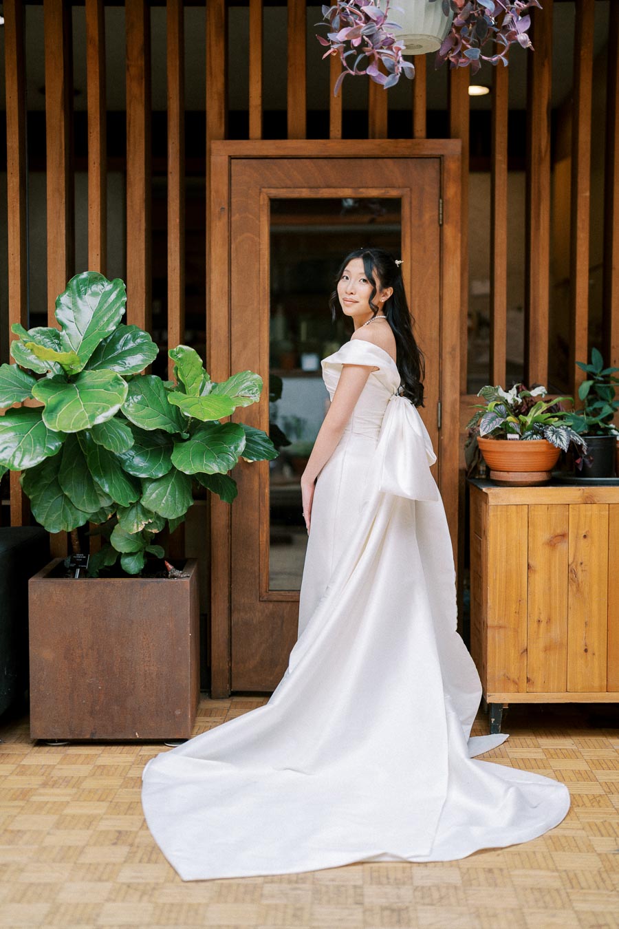 A bride in an elegant off-shoulder white wedding gown poses in front of a wooden door, surrounded by lush green potted plants indoors.