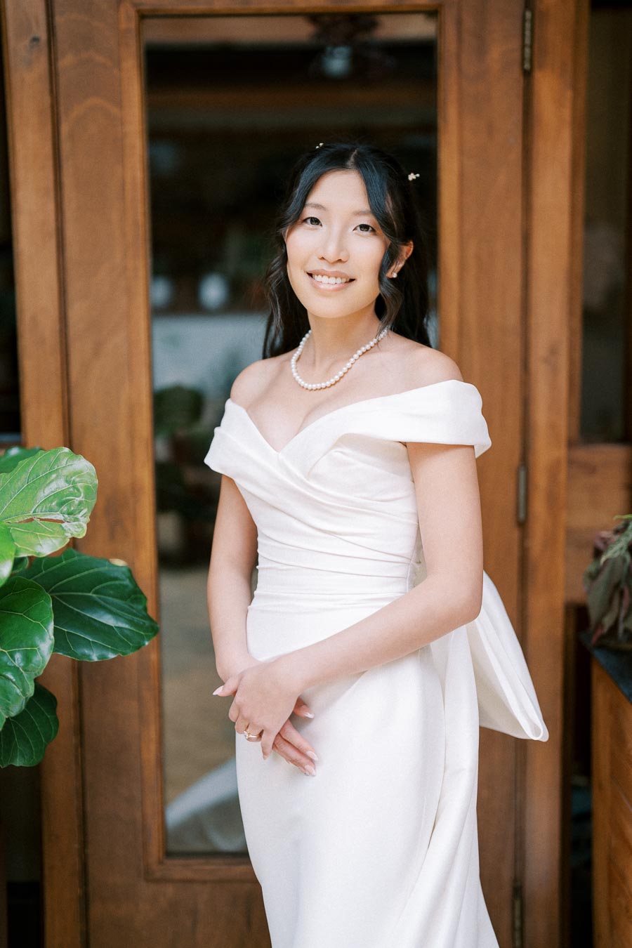 A bride in an elegant white dress poses in front of a wooden door, wearing a pearl necklace and smiling softly, with lush green plants on either side.