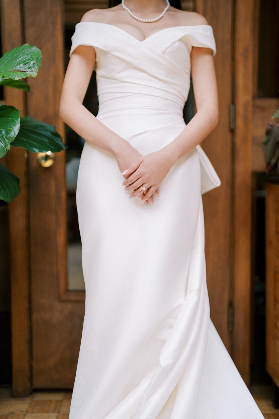 Elegant bride in an off-the-shoulder white wedding dress, standing with hands clasped in front, adorned with a pearl necklace, against a wooden door background.