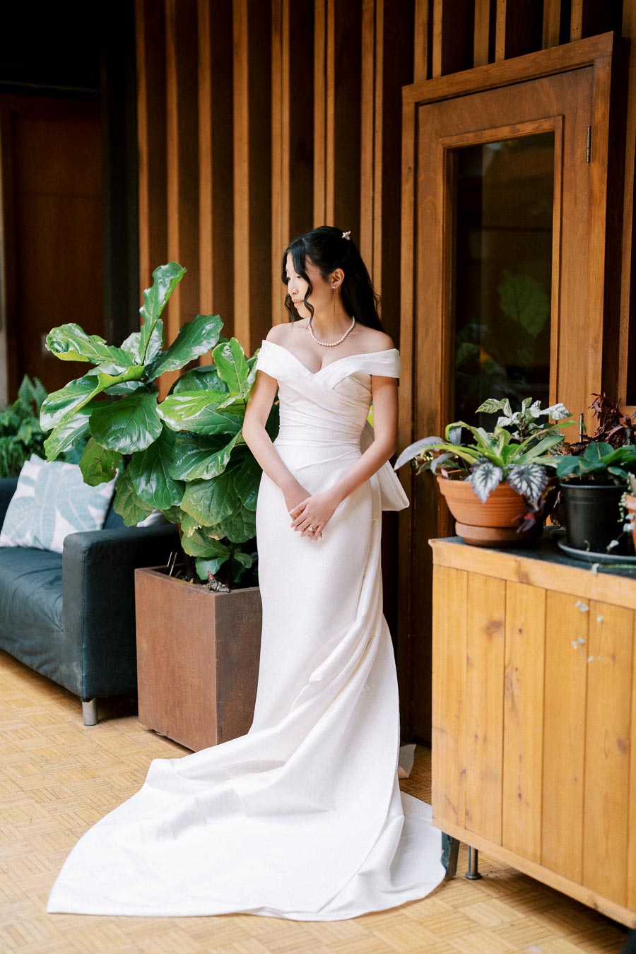 A bride in an elegant off-the-shoulder white wedding gown stands inside a stylish room with wooden walls, surrounded by lush green plants.