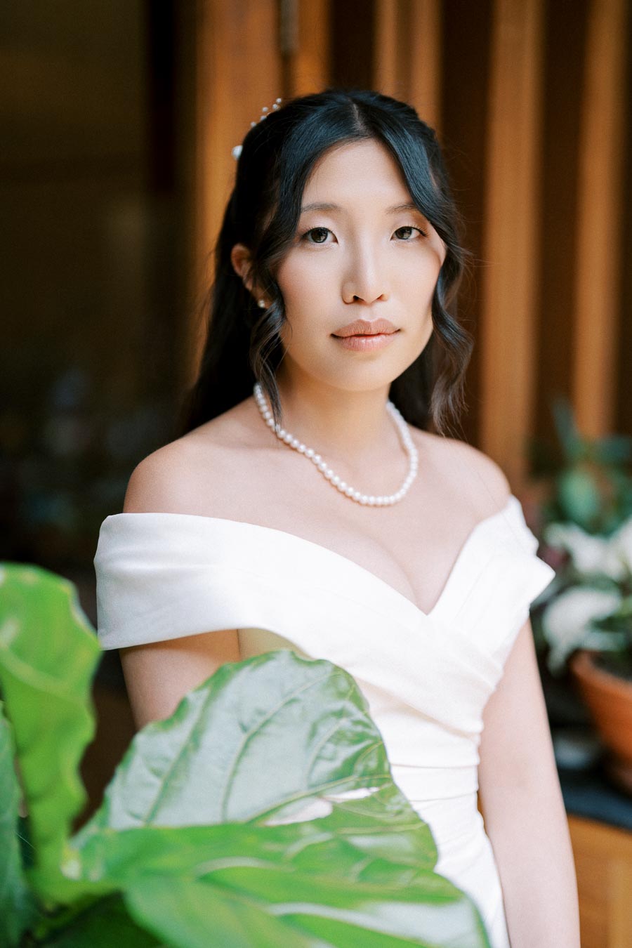 Elegant woman in an off-shoulder white dress with pearl necklace, standing near green foliage indoors.