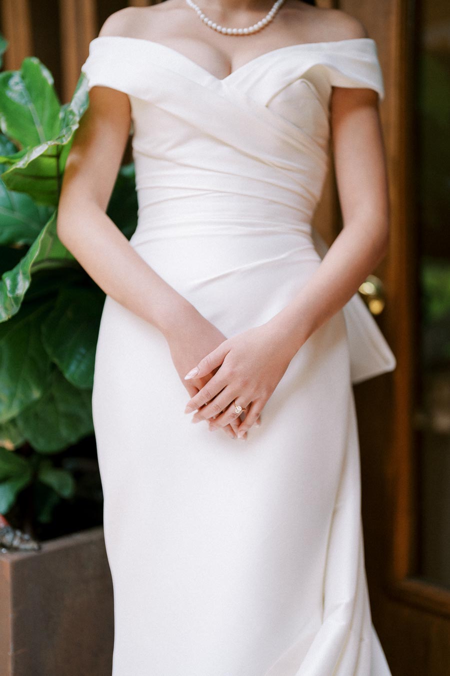 A bride in an elegant white off-shoulder wedding dress adorned with a pearl necklace and a diamond ring, standing near lush green plants.