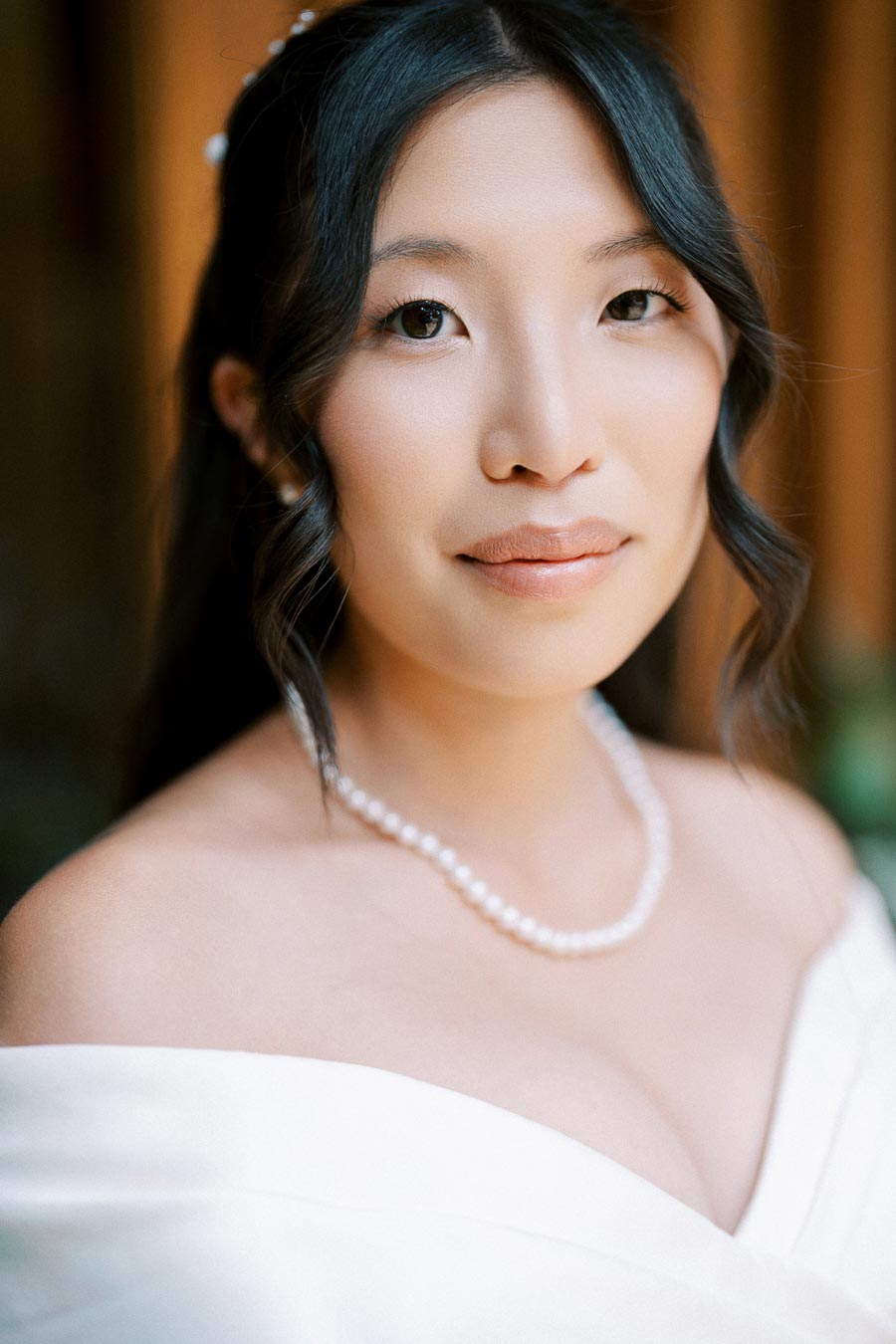 A close-up portrait of a bride in a white off-shoulder gown, wearing a pearl necklace with softly curled hair and a serene expression.