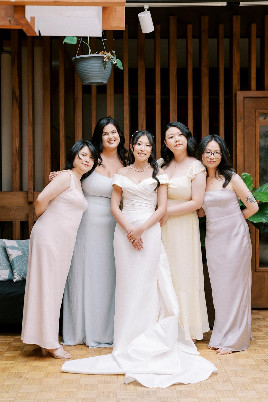 Smiling bride in elegant white gown with bridesmaids in pastel dresses, posing at a wedding ceremony with wooden background and hanging plant decor.