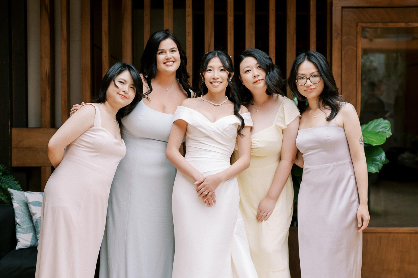 A group of five women in elegant pastel dresses posing together, with one woman in a white bridal gown at the center, standing in front of a wooden backdrop with greenery, suggesting a wedding celebration or bridesmaid gathering.