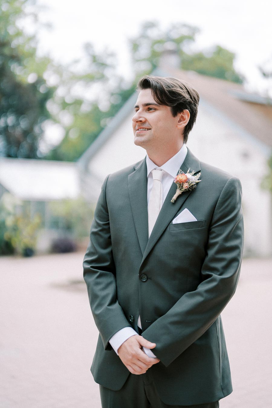 Man in a dark suit smiling outdoors at a wedding venue, wearing a boutonniere and standing against a blurred background of greenery and architecture.
