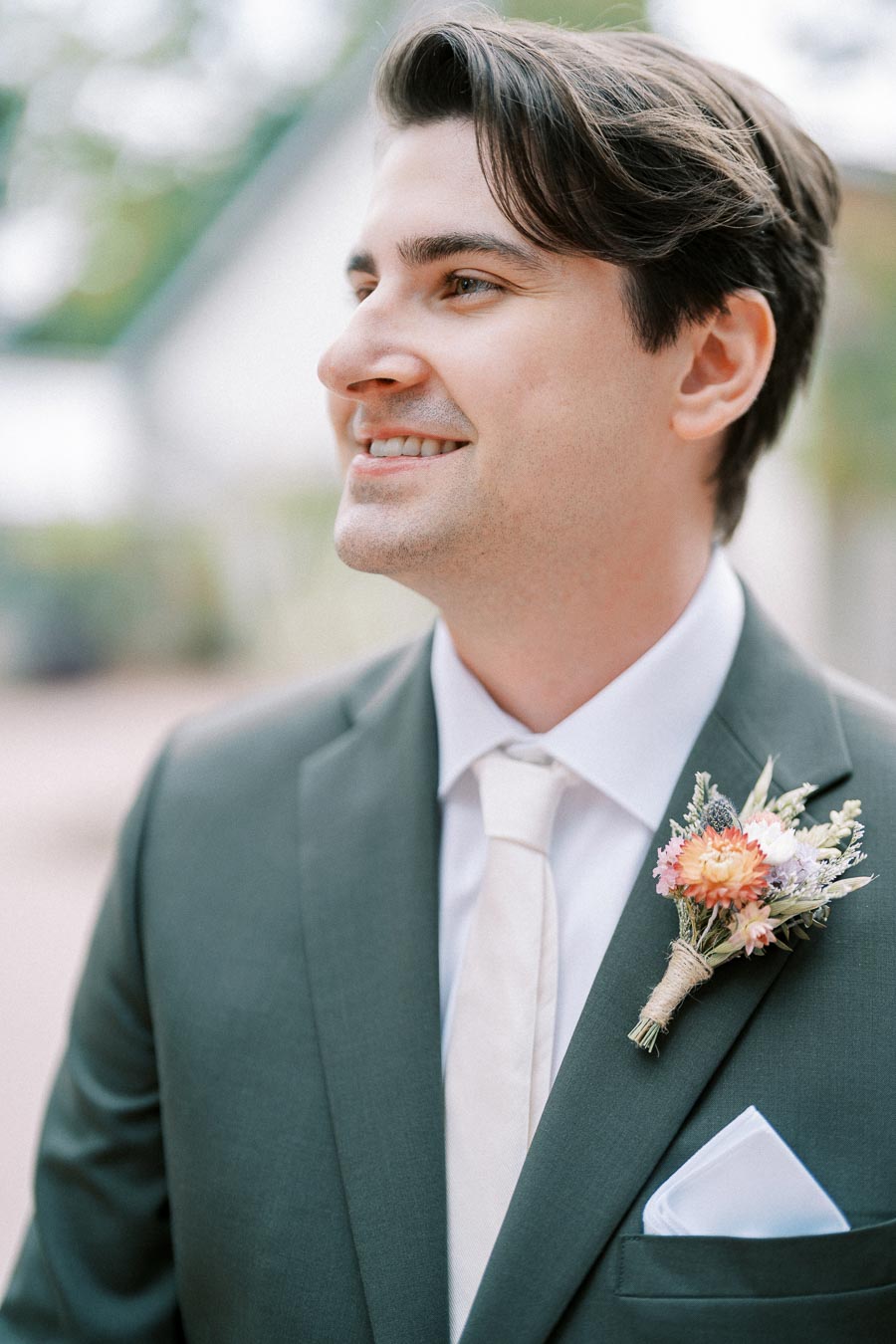 A groom wearing a dark suit and white tie with a colorful floral boutonniere, smiling on his wedding day.