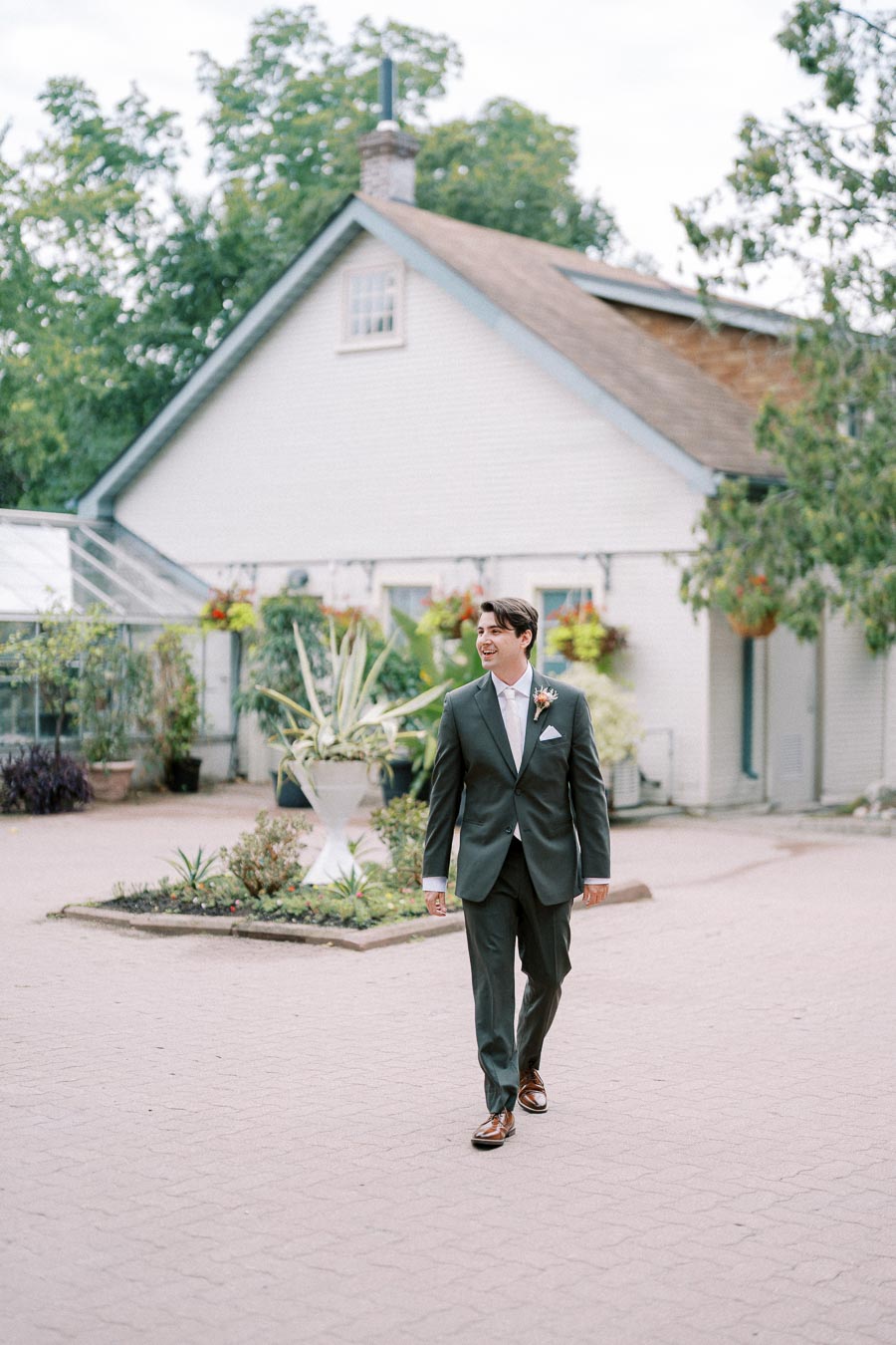 A man in a suit walks confidently in a garden courtyard in front of a rustic building, surrounded by greenery and potted plants.