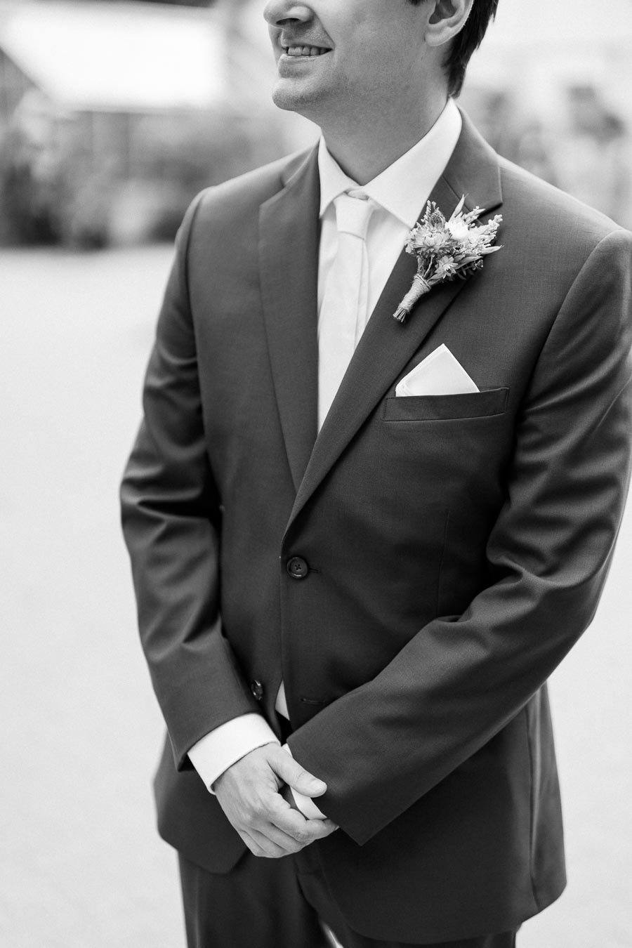 Black and white image of a groom in a suit with a boutonniere, standing and smiling, hands clasped in front.