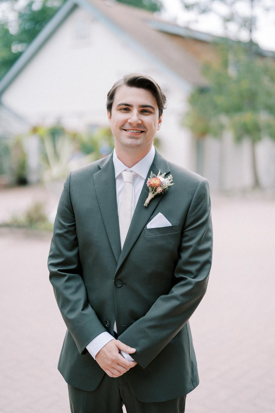 A groom in a dark suit with a boutonniere smiles while standing outdoors in front of a blurred background with greenery and a house.