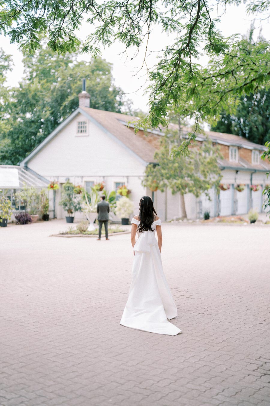 Bride in white wedding dress approaches groom in courtyard surrounded by greenery and charming buildings, captured during a romantic outdoor wedding photoshoot.