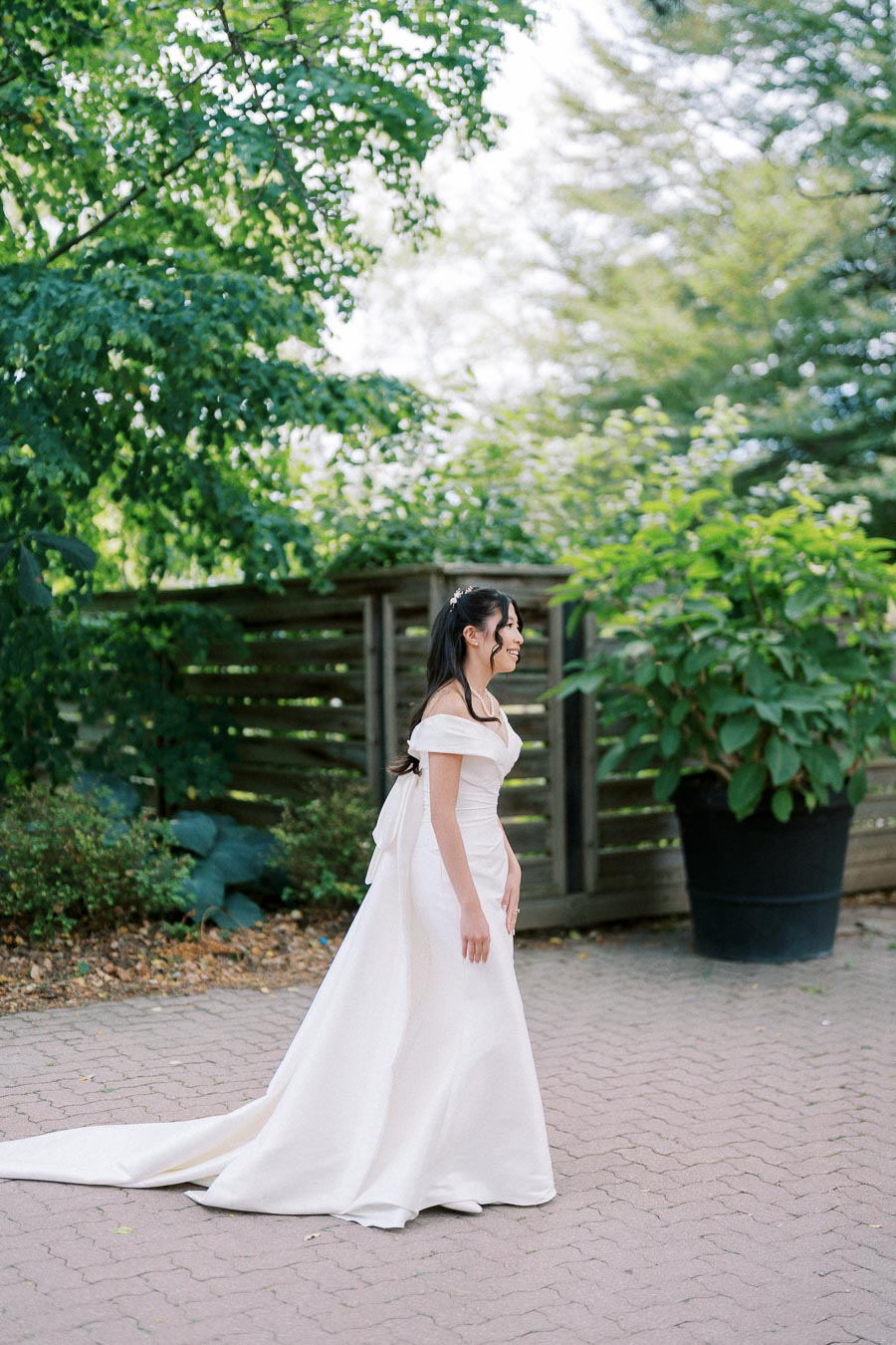 Bride in a flowing white gown smiles while walking outdoors, surrounded by lush greenery and wooden fencing.