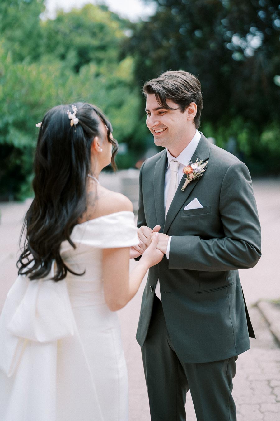 A bride and groom holding hands and smiling at each other in an outdoor setting, with greenery in the background, on their wedding day.