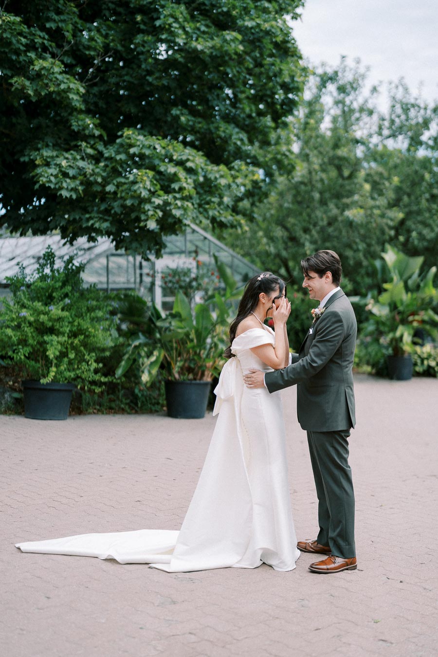 A bride in an elegant white gown shares an emotional first look with her groom, dressed in a stylish suit, in a lush garden setting with greenery and a greenhouse in the background.