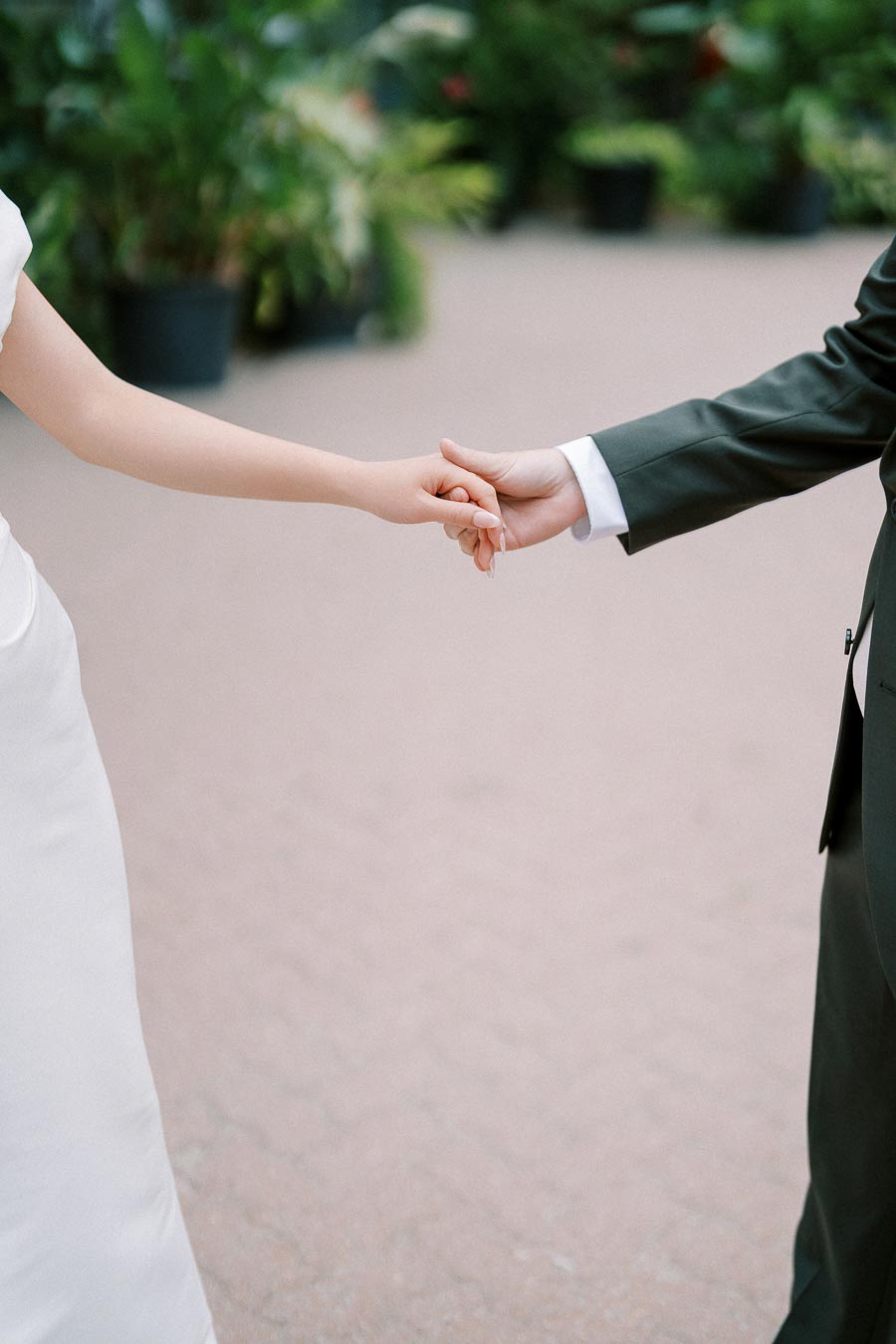 A close-up of a couple holding hands, with one person wearing a white dress and the other in a dark suit, set against a blurred outdoor background with greenery.