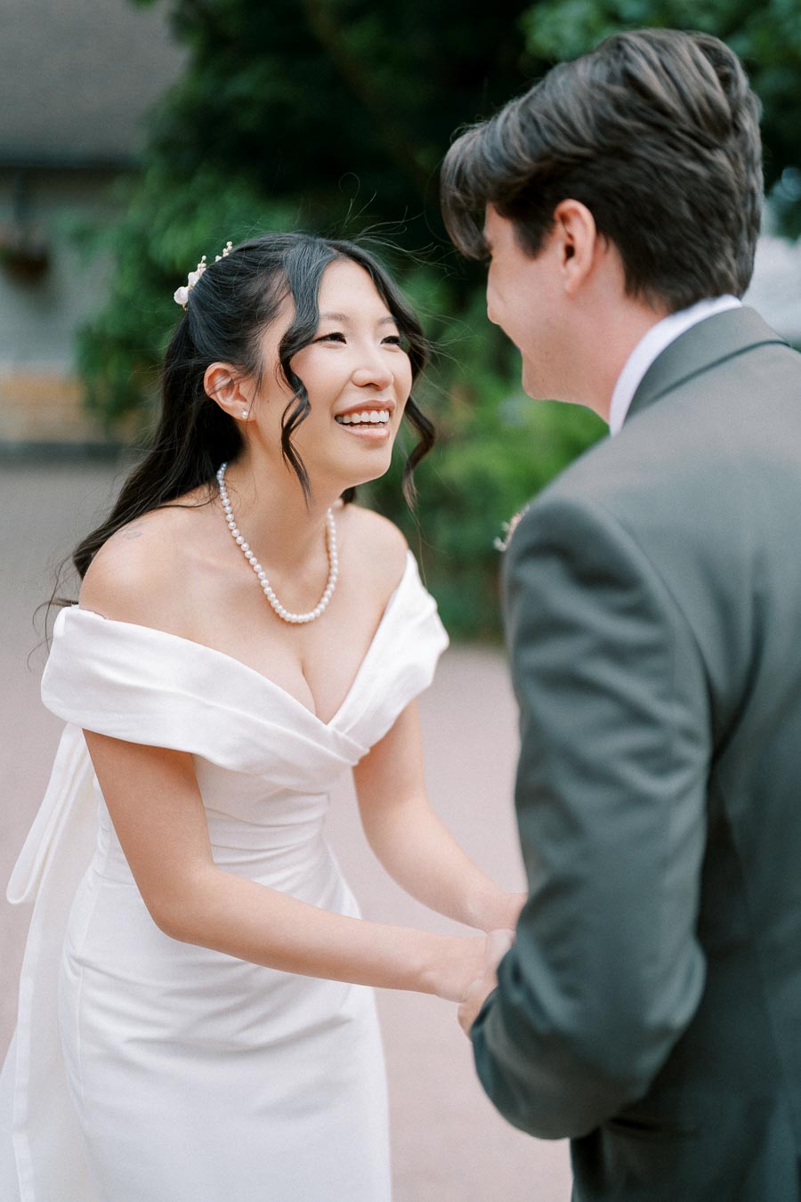 A smiling bride in an elegant white wedding dress shares a joyful moment with the groom in a gray suit outdoors, surrounded by greenery.