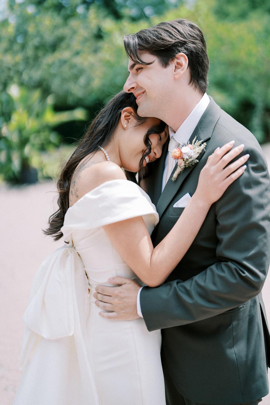 A bride and groom lovingly embrace outdoors on their wedding day, with the bride in an elegant white dress and the groom in a classic suit with a floral boutonnière, set against a lush green background.