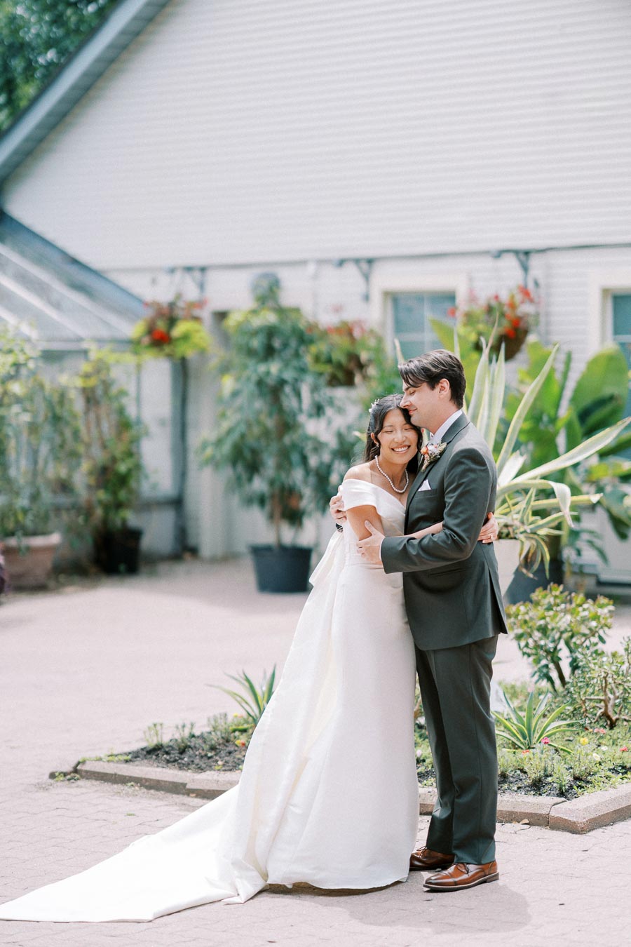 A happy couple embracing on their wedding day outside a charming white building, surrounded by lush greenery and decorative potted plants.