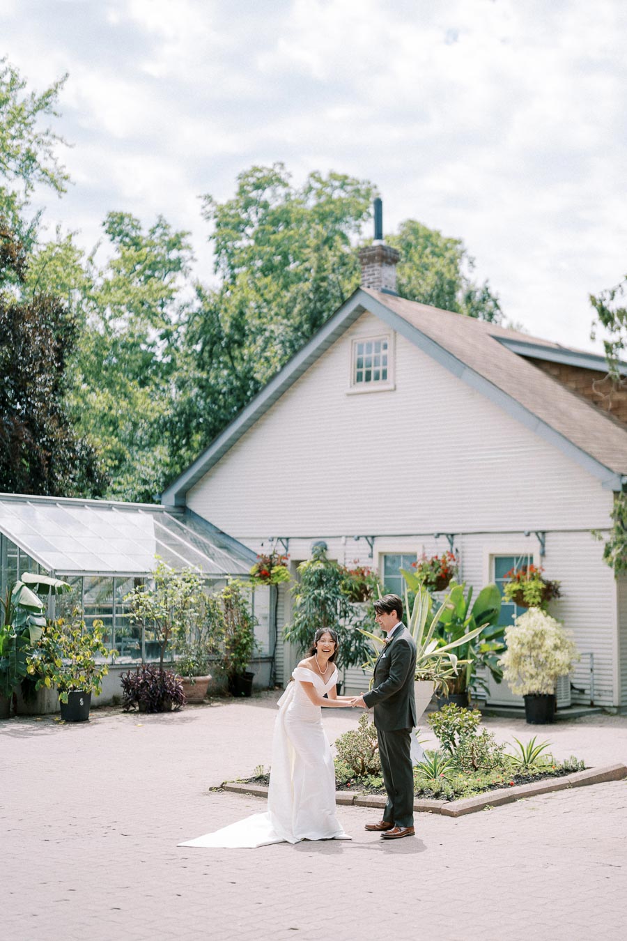 A bride and groom laughing joyfully in a garden courtyard, surrounded by lush greenery and a charming white cottage, creating a picturesque wedding scene on a sunny day.