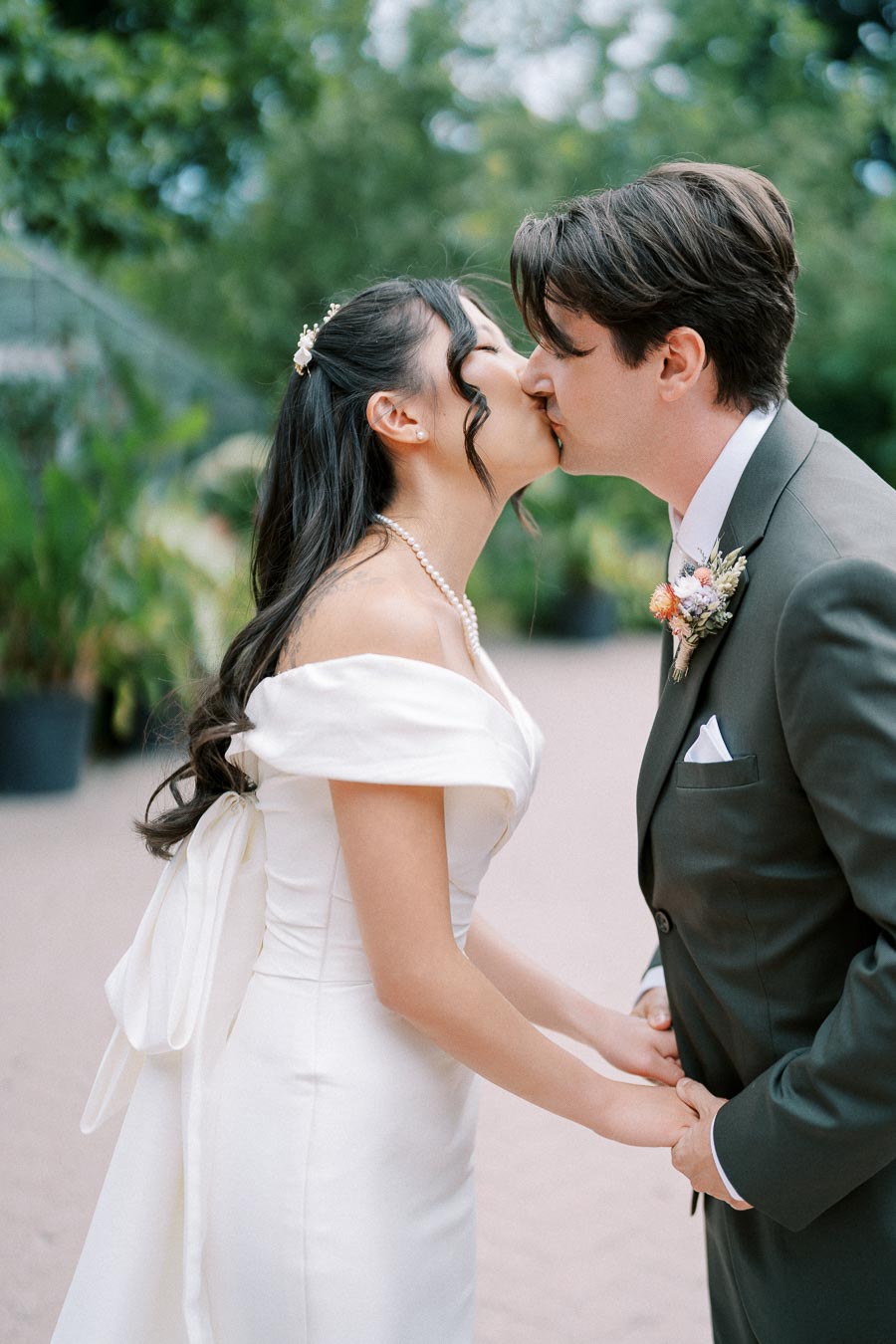 Bride and groom sharing a kiss outdoors, bride in elegant white dress with pearl necklace, groom in dark suit with boutonniere.
