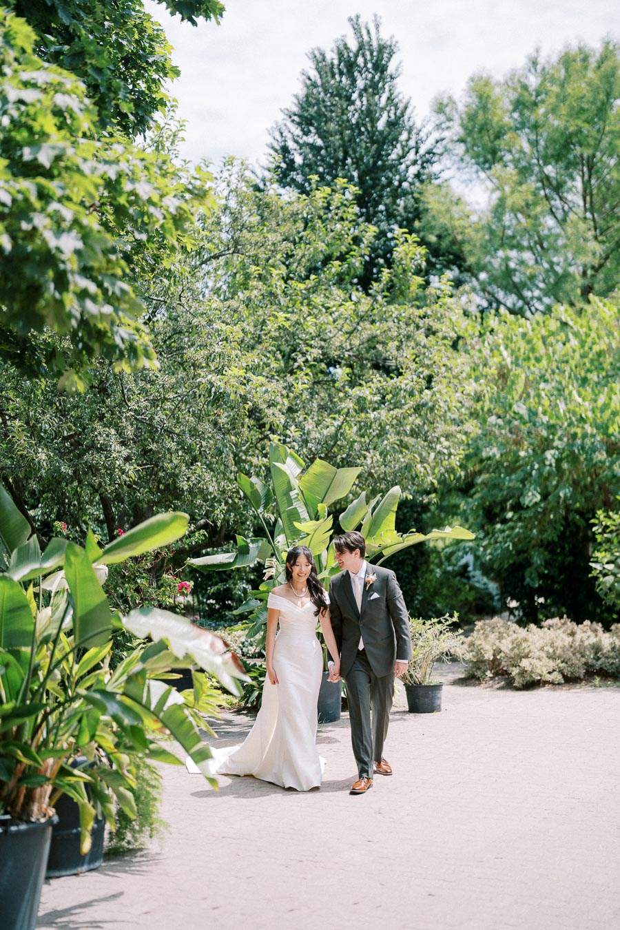 Wedding couple walking hand in hand through lush garden surrounded by greenery and plants on a sunny day.