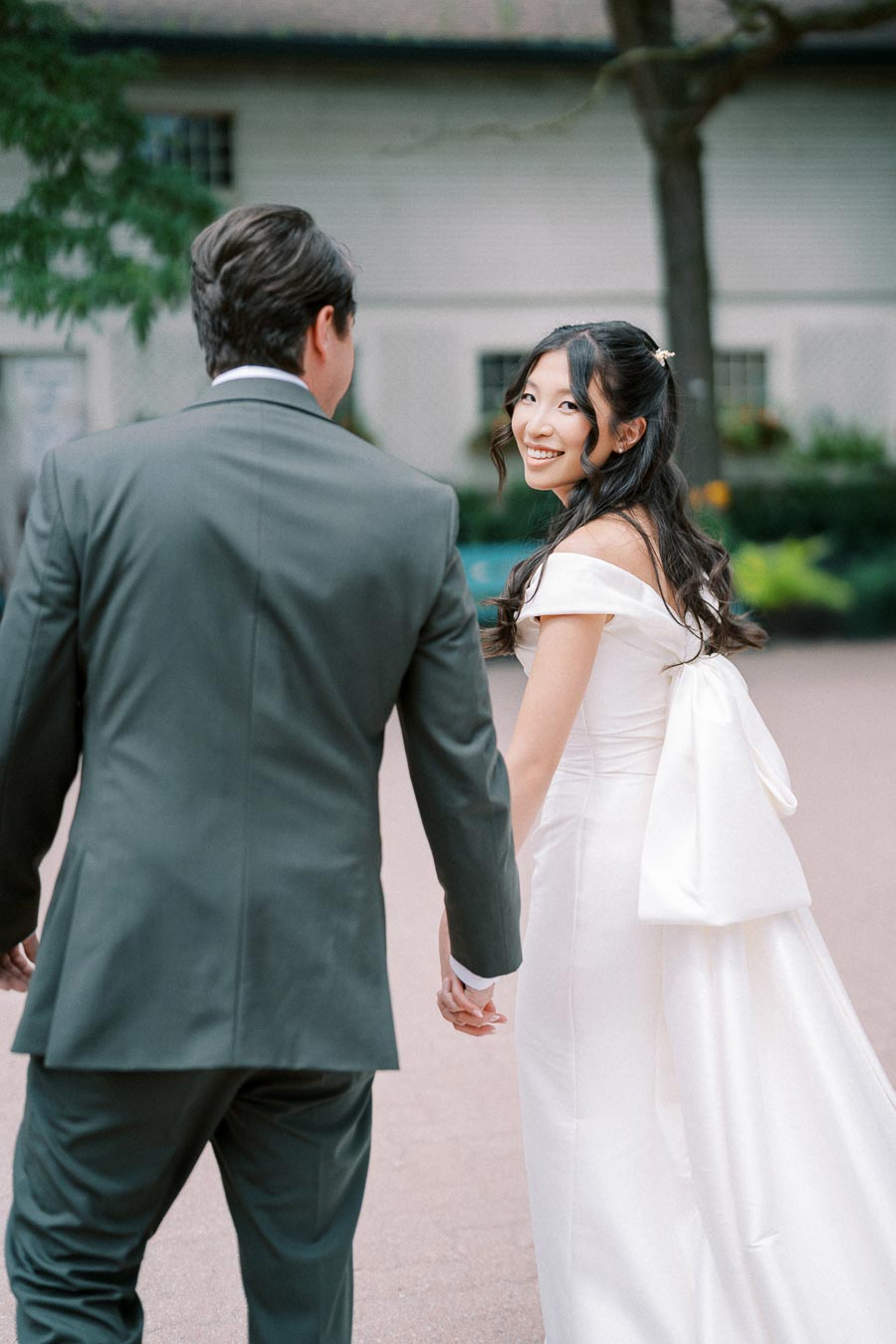 Bride and groom hold hands, smiling at each other in an outdoor setting, with the bride in a white gown and the groom in a dark suit.