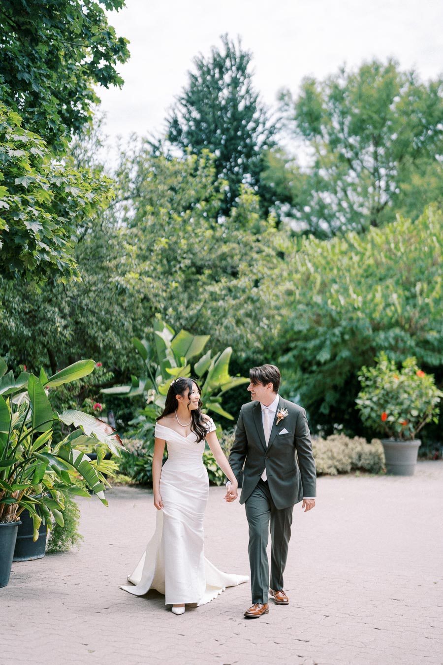 A bride in an elegant white gown and a groom in a classic black suit walking hand-in-hand through a lush garden on a sunny day, showcasing a beautiful outdoor wedding scene.