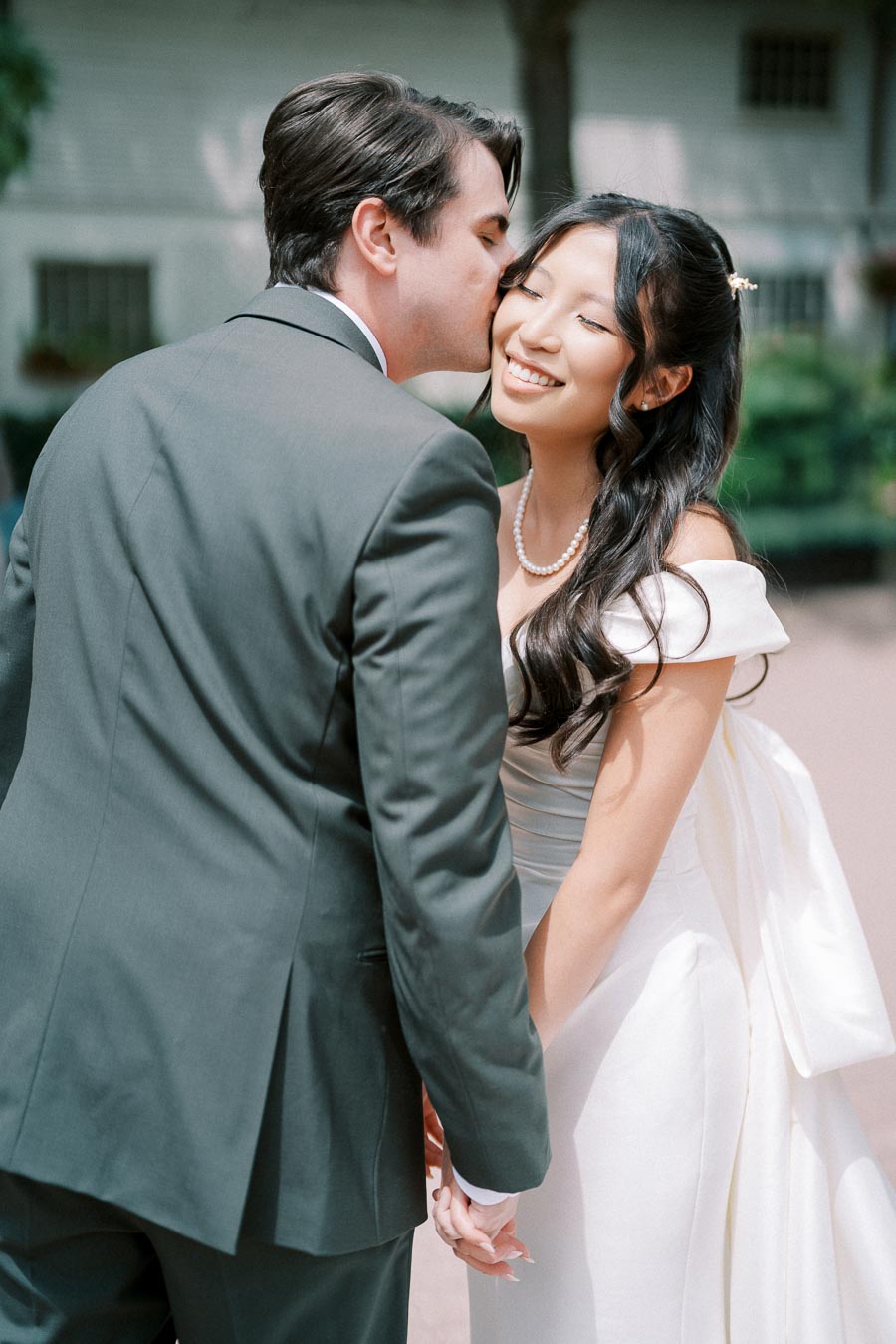 A groom in a dark suit kisses the smiling bride in a white wedding dress, holding hands outdoors.