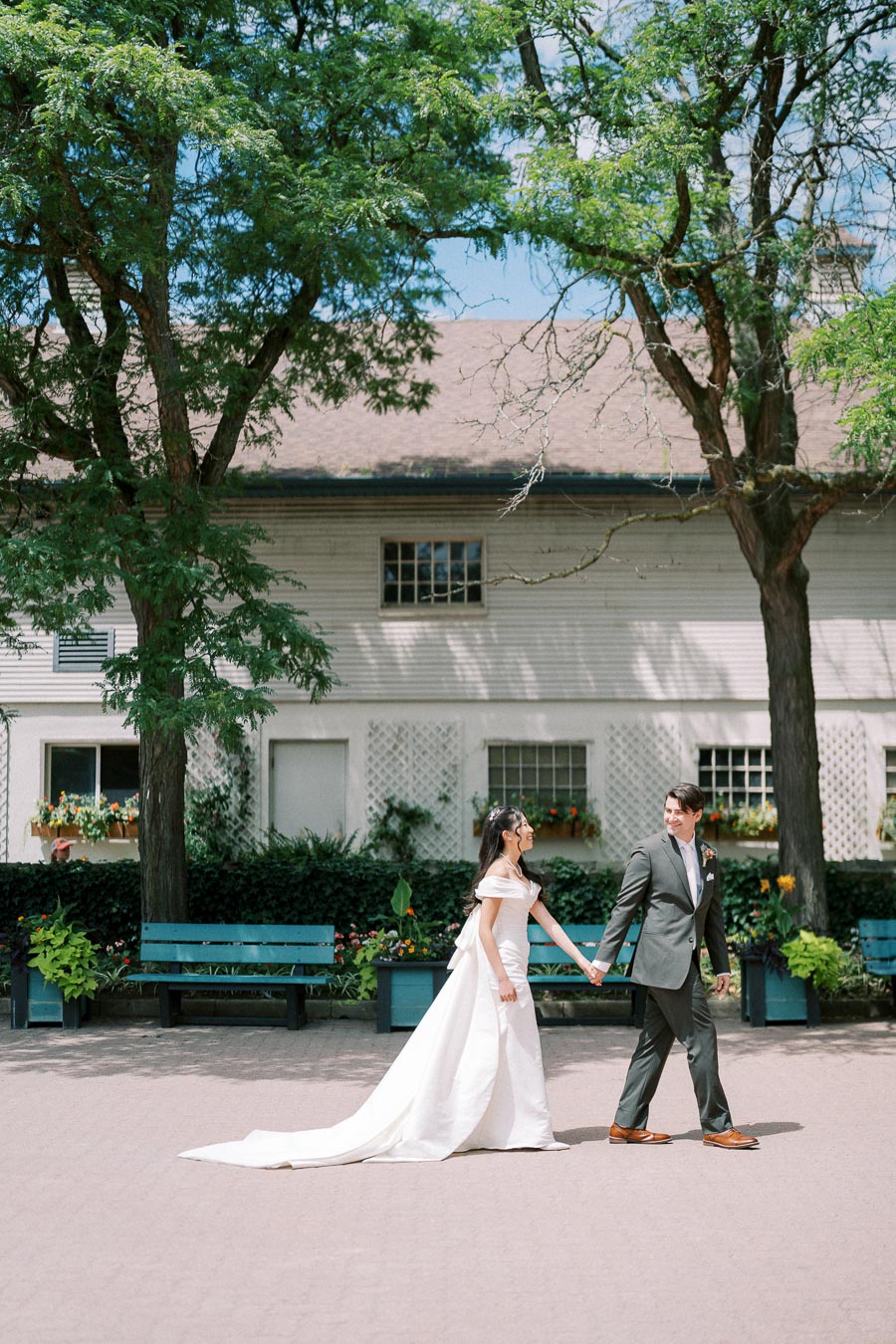 Wedding couple holding hands and walking in a charming outdoor setting with lush greenery and a quaint building in the background.
