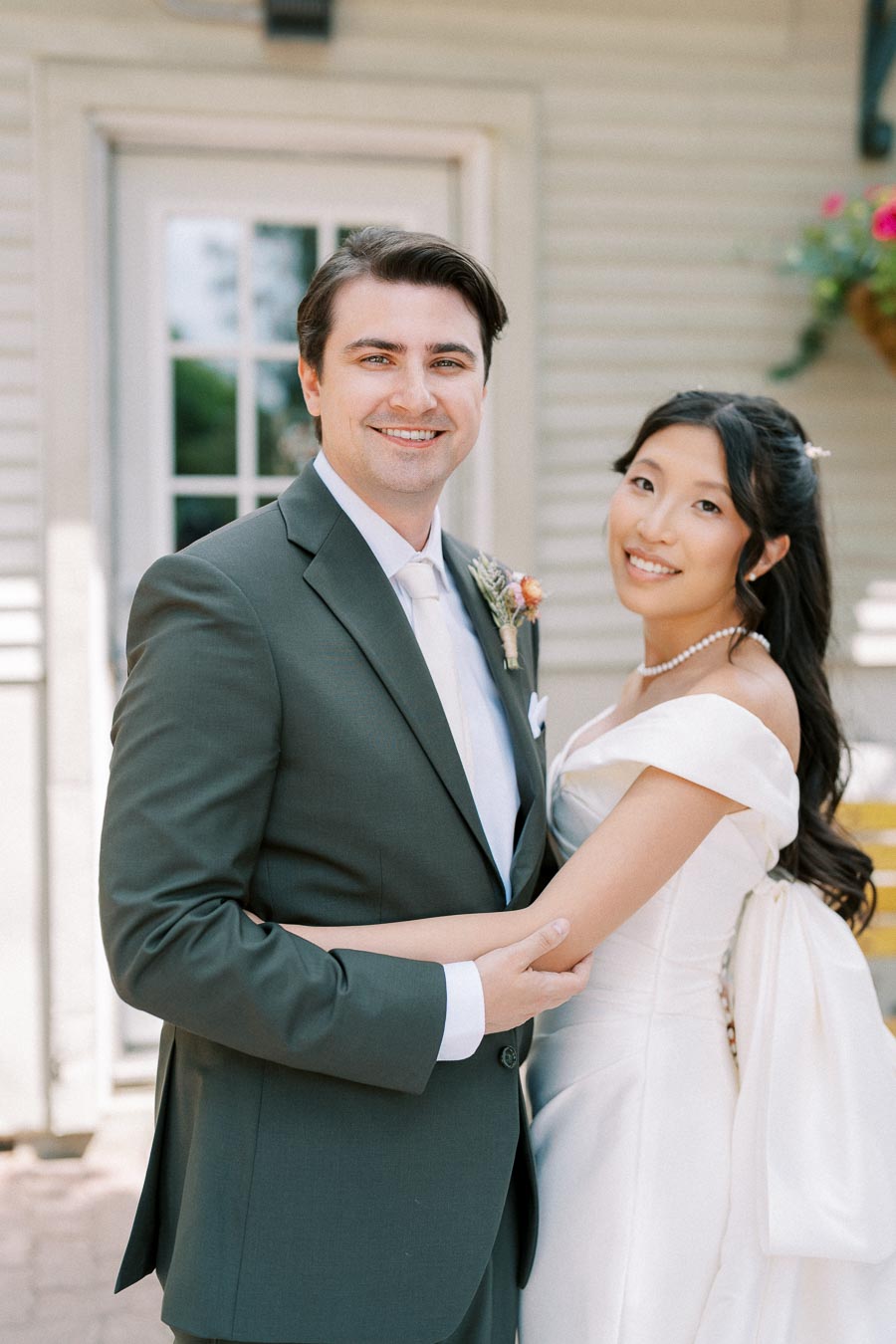 A happy couple in wedding attire, with the groom in a dark suit and the bride in a white gown, smiling and embracing in front of a house with flowers.
