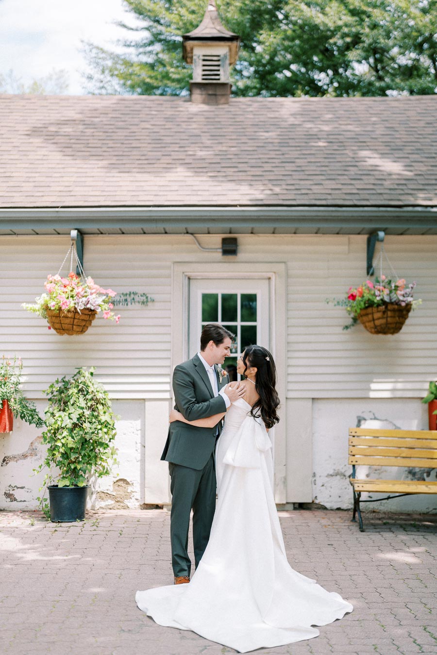 A bride in a flowing white gown and groom in a gray suit embrace in front of a charming cottage with hanging flower baskets and lush greenery.