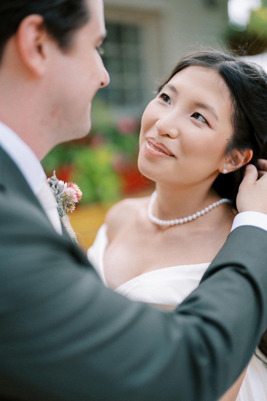 A bride and groom share an intimate moment on their wedding day, with the bride gazing lovingly at the groom. She is wearing a white dress and pearl necklace, while the groom is dressed in a suit, holding a small bouquet. The background features soft, colorful floral decorations, creating a romantic atmosphere.