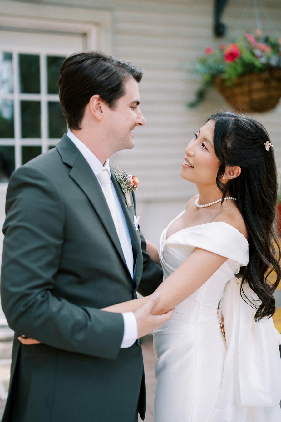 A bride and groom lovingly gaze at each other during their outdoor wedding ceremony, with the bride in an elegant white gown and the groom in a black suit, standing in front of a house with a flower basket in the background.