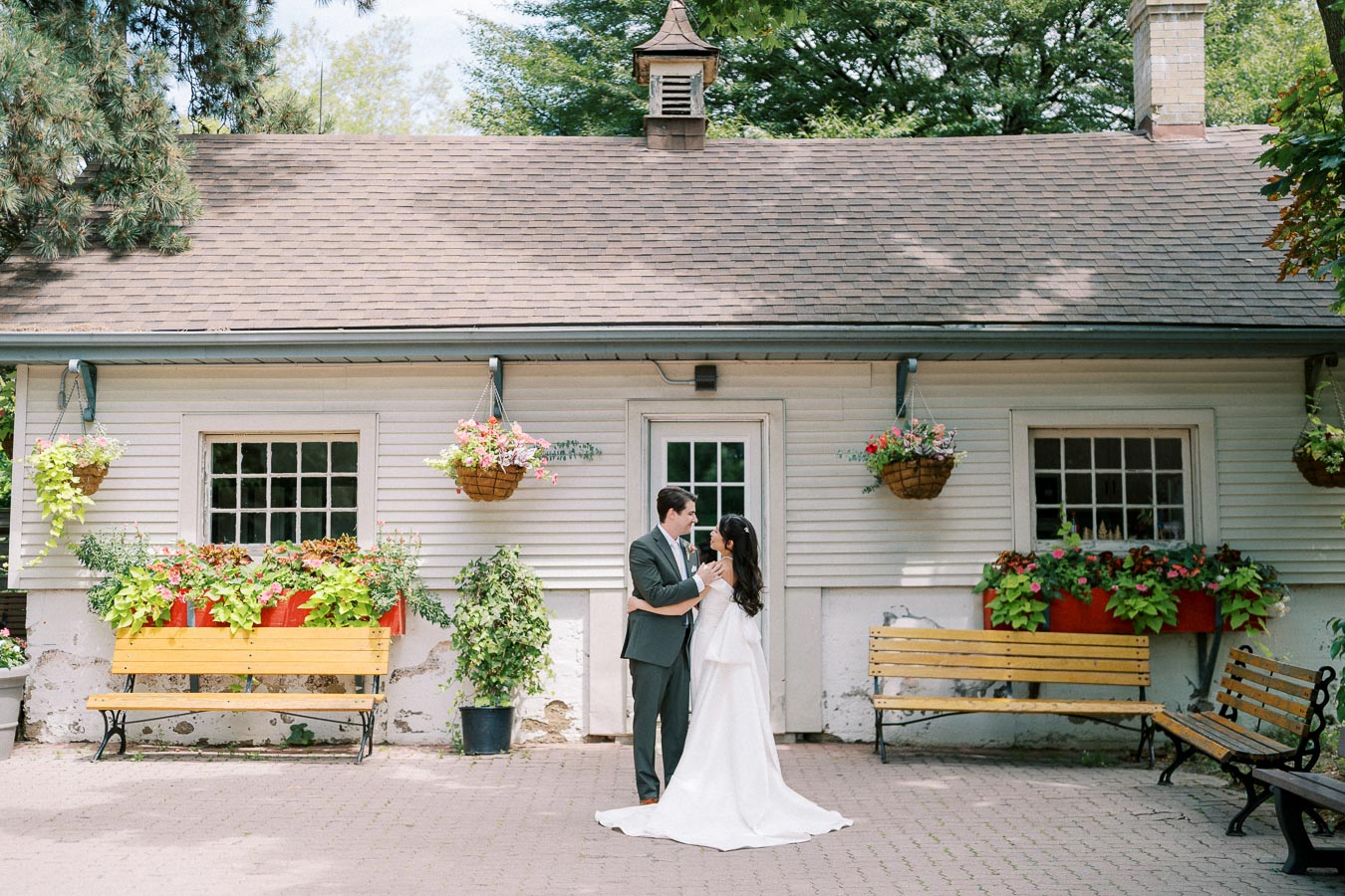 A bride and groom embrace in front of a charming cottage with hanging flower baskets and wooden benches, creating a picturesque outdoor wedding scene.