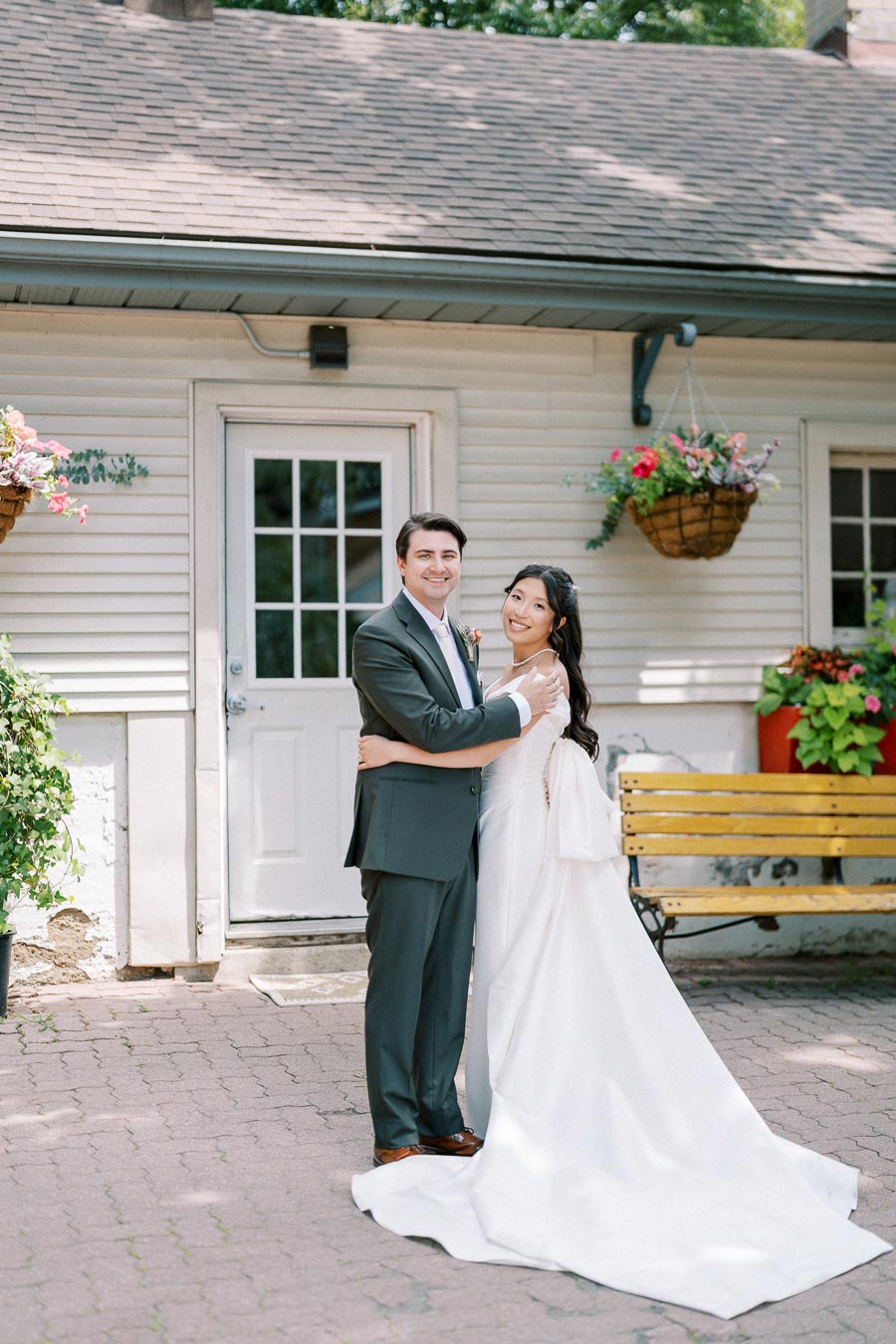 A beaming couple in elegant wedding attire embraces in front of a charming house with a wooden bench and vibrant hanging flower baskets.