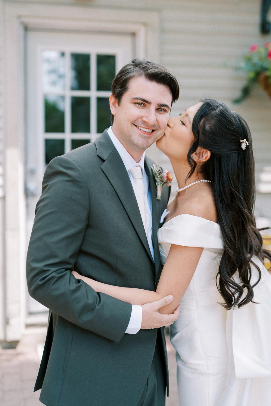 Wedding couple embracing, bride in white dress kissing groom in suit outdoors on a sunny day, showcasing love and romance.