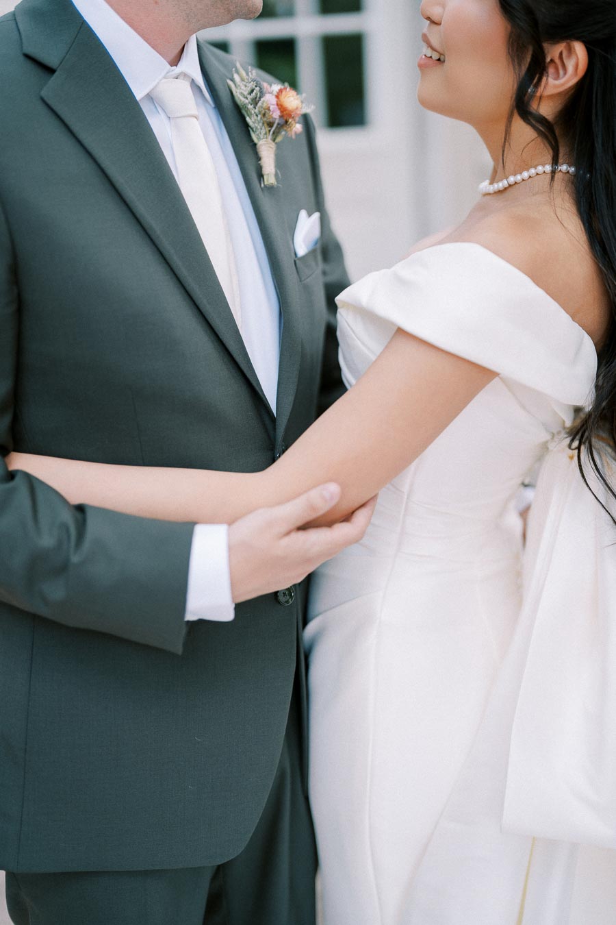 Bride and groom embracing on their wedding day, with the bride in an elegant white off-shoulder dress and pearl necklace, and the groom in a sharp dark suit with a boutonnière.