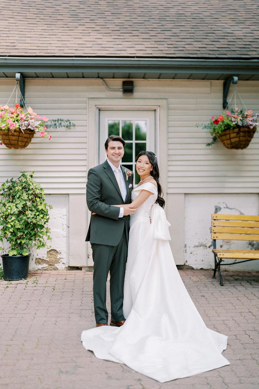Wedding couple standing in front of a rustic building with hanging baskets and a bench, bride in elegant white gown, groom in dark suit, both smiling.
