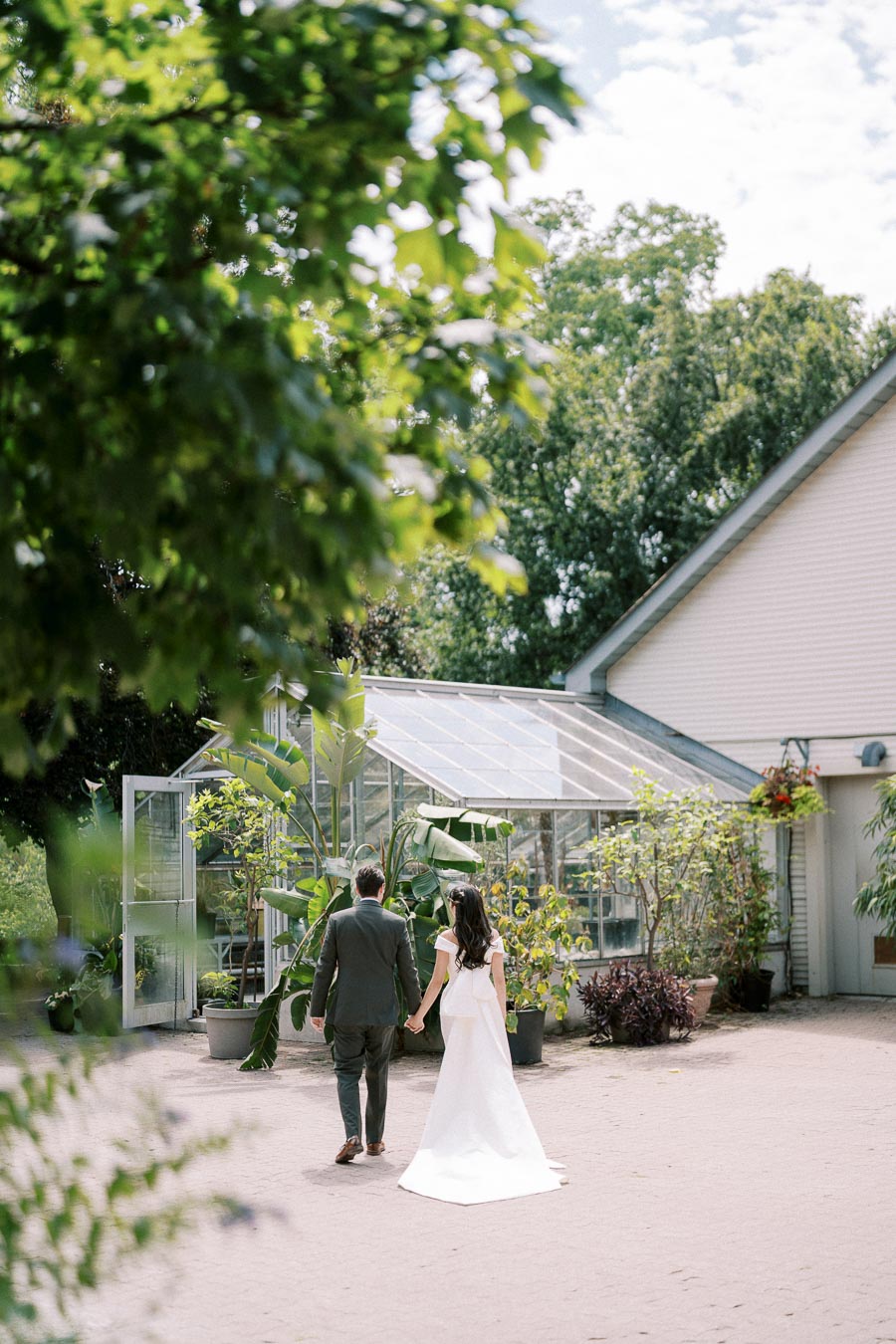 Wedding couple holding hands walking towards a glass greenhouse, surrounded by lush greenery and trees, under a clear blue sky.